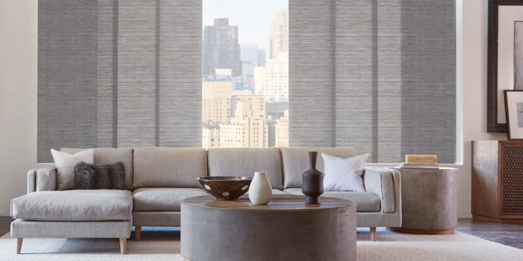 A living room with a grey couch and blinds for large windows, offering a view of the city.