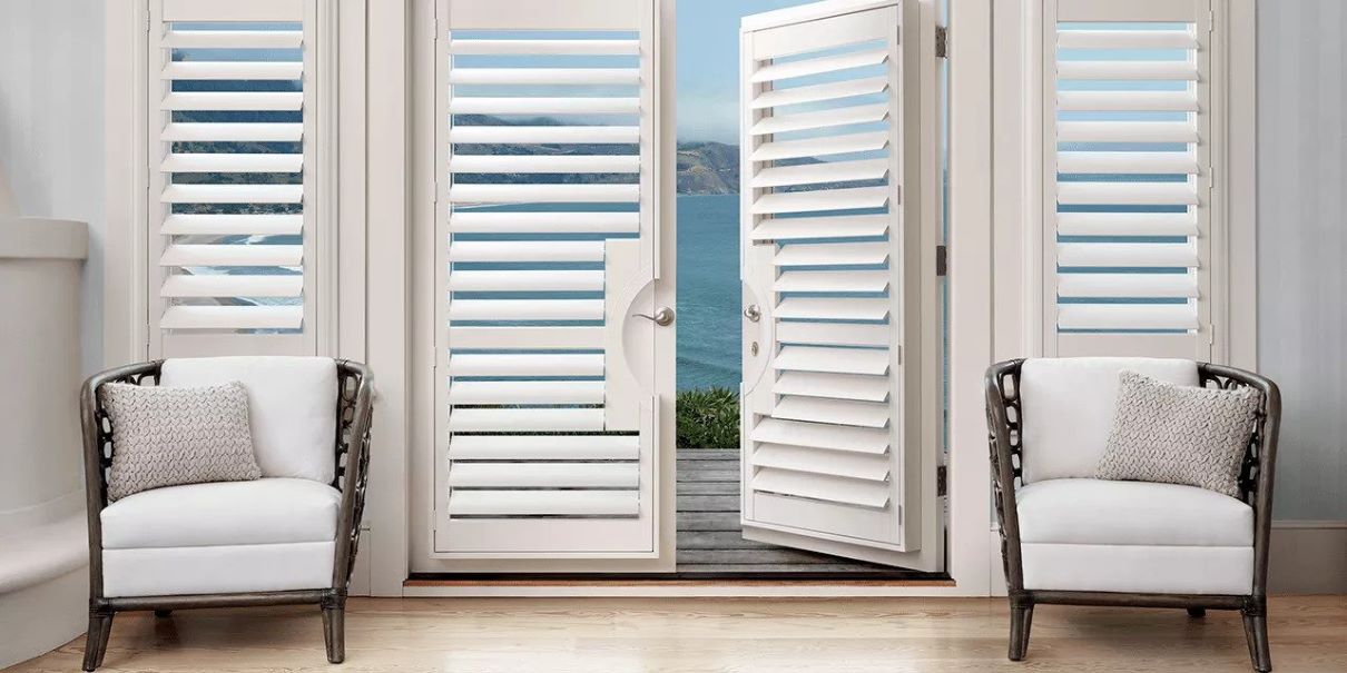 A room with white shutters and a view of the ocean through its front door window coverings.