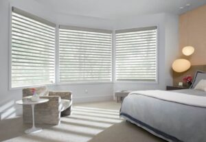 A bedroom with large windows fitted with sheer shades and a comfortable bed.